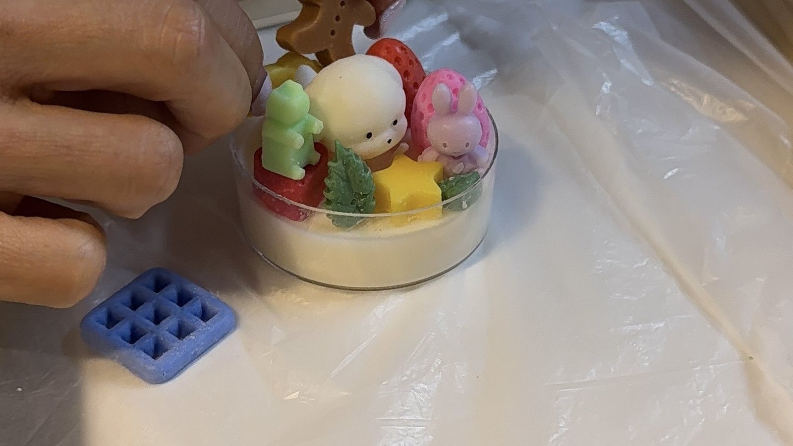 A hand holds a candle decorated with a bunny, flowers and a strawberry beside a wax mould
