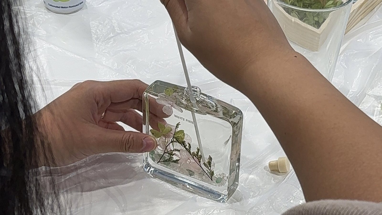 A hand uses tweezers to place dried flowers into a glass diffuser bottle