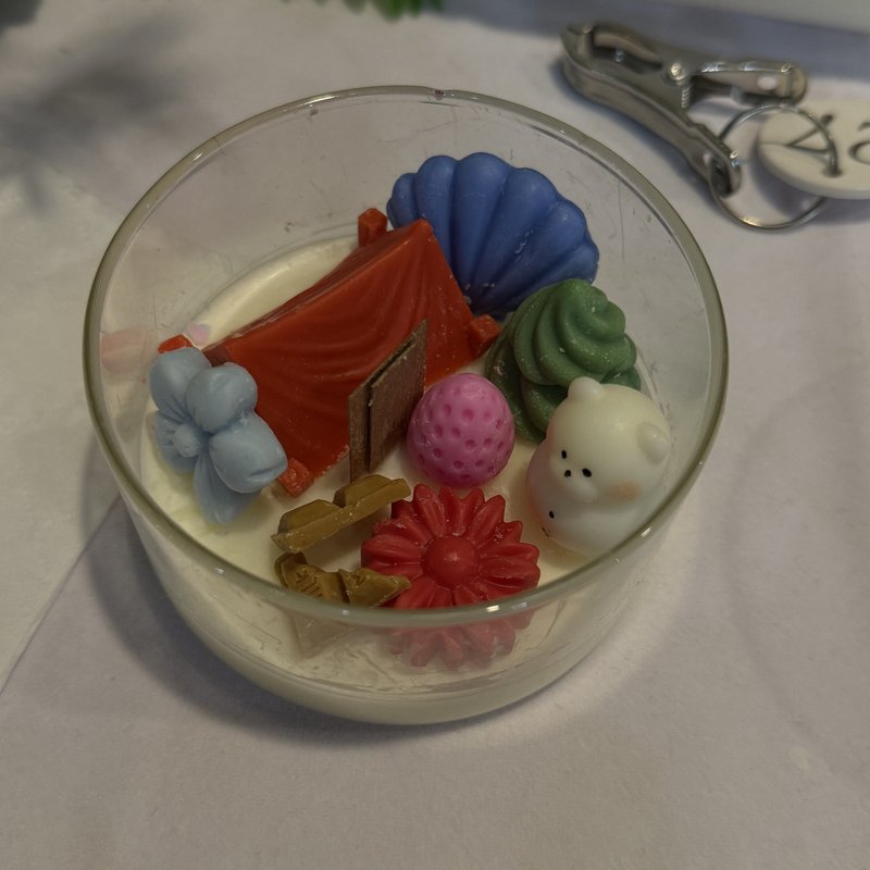 A candle in a glass bowl decorated with wax flowers, a strawberry and a small wax bear