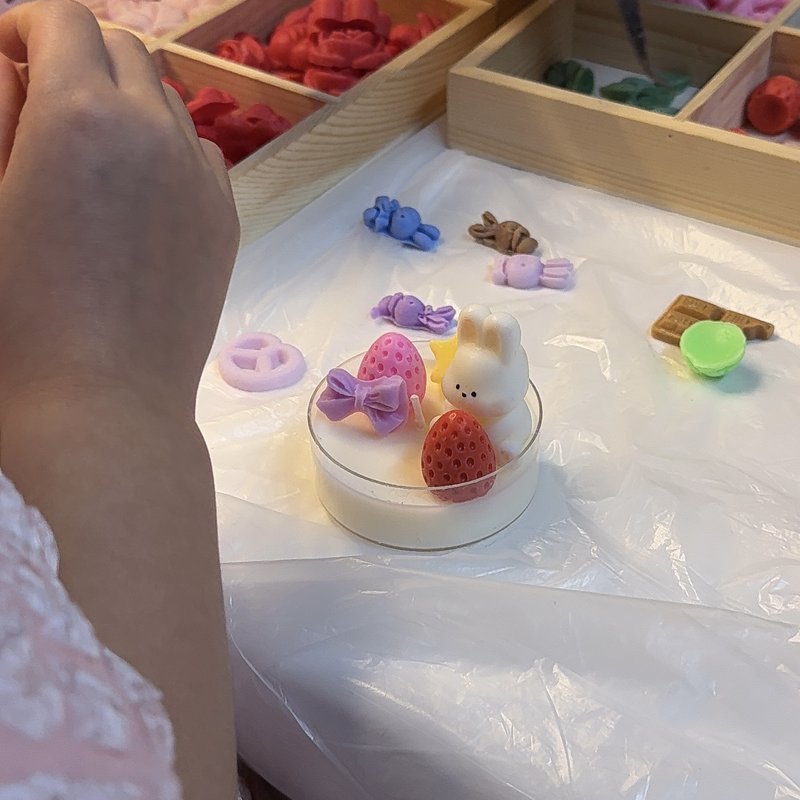 A small candle decorated with a wax bunny, strawberries and sweets on a white workshop table