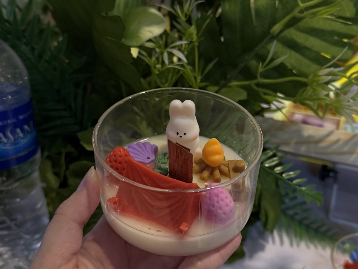 A candle in a glass cup with a small bunny figure, wax flowers and greenery behind it