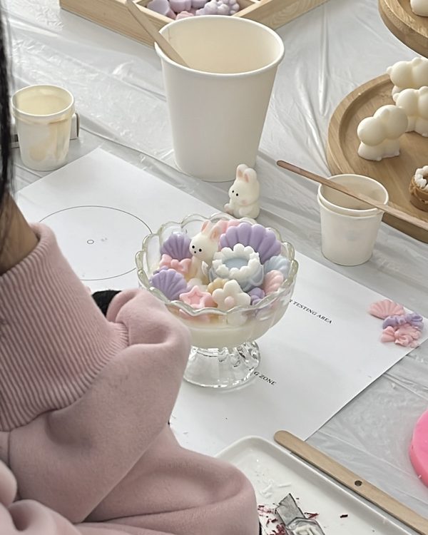 A first candle in a glass cup, topped with pastel wax flowers and a small wax bunny, on a workshop table
