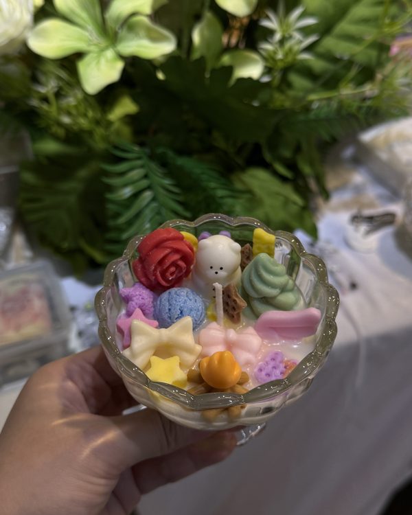 A candle in a glass cup decorated with wax roses, a small bear and pastel sweets