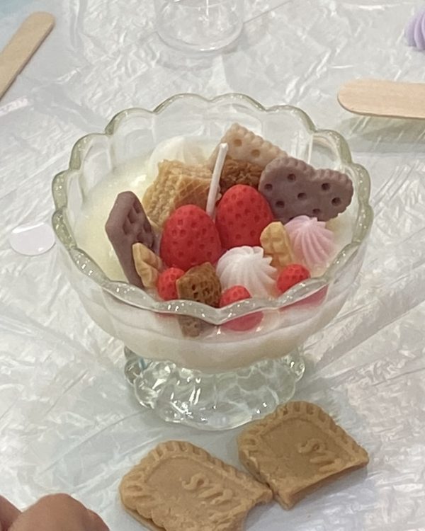 A candle in a glass cup filled with wax strawberries, hearts, cookies and whipped wax swirls